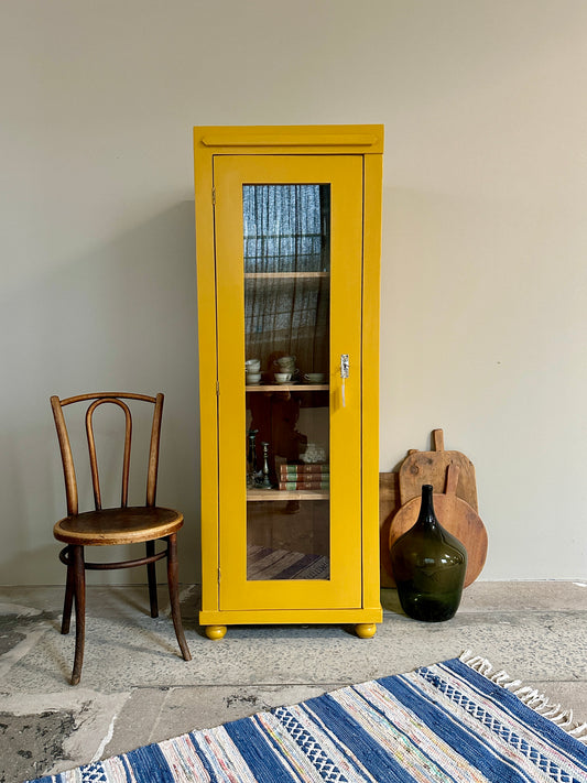 Bild visar ett ockragult vitrinskåp med 1 dörr samt en träfärgad stol