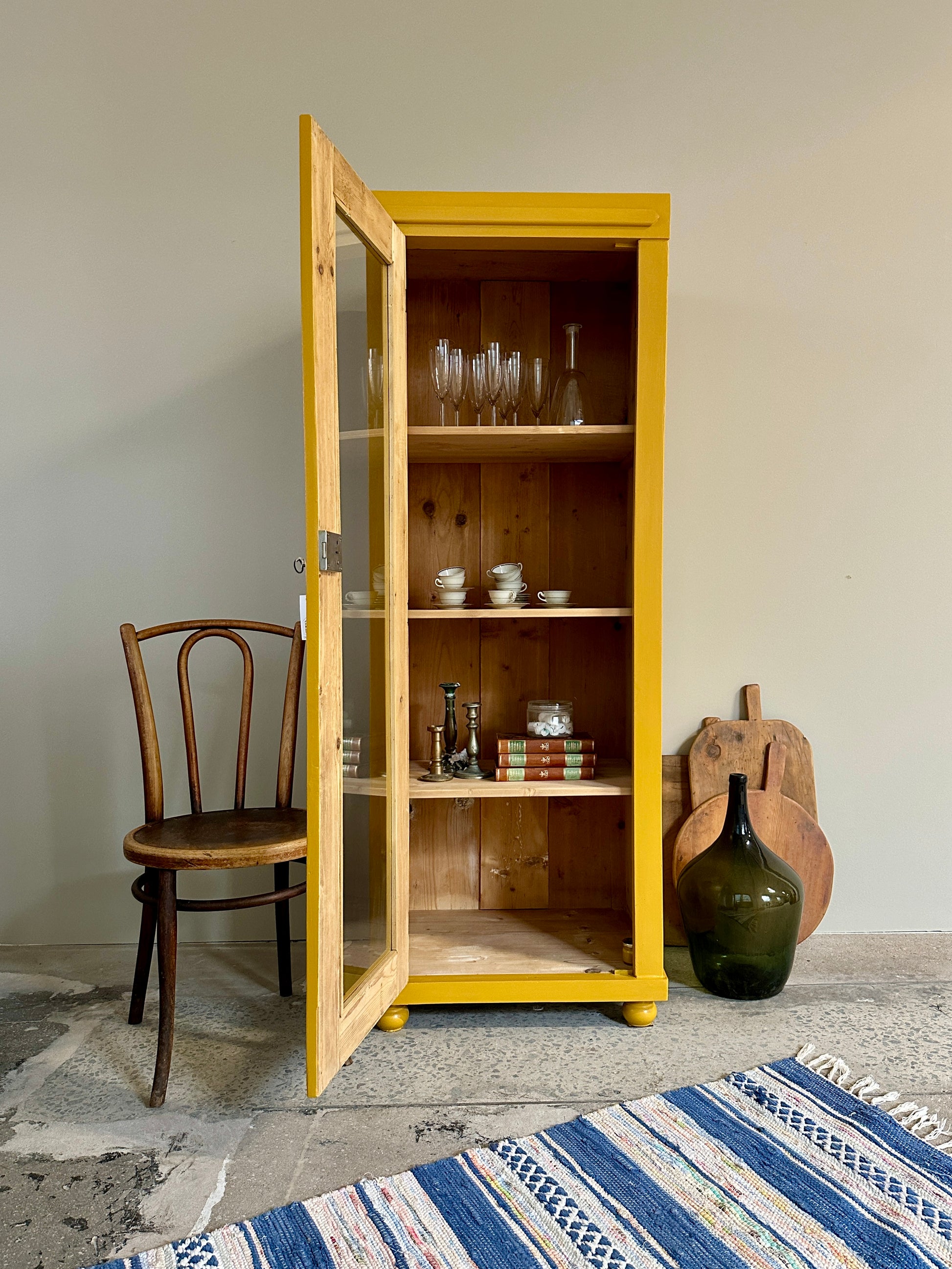 Bild visar ett ockragult vitrinskåp med 1 dörr samt en träfärgad stol. Bild visar med öppen dörr