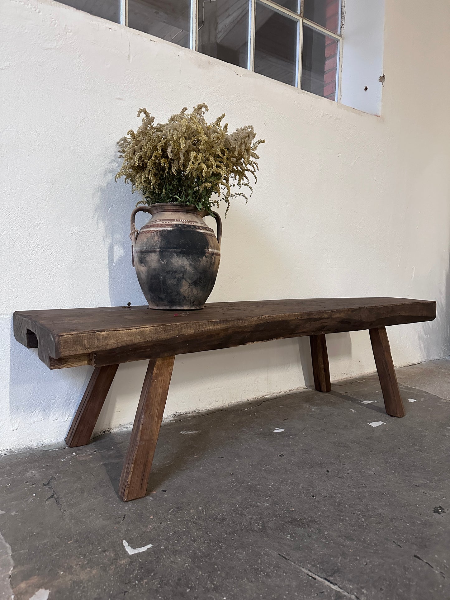 Träbänk mot vit vägg. Uppe på bänken står en urna med torkade blommor i. Bilden är tagen från sidan.