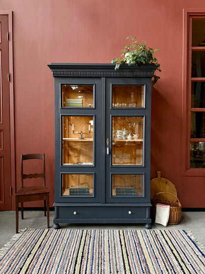 Ett gammalt, otroligt vackert vitrinskåp med fina detaljer i de raka formerna.Skåpet har i dagsläget 3 st nya och fräscha hyllplan, vi kan sätta in fler om det önskas. Det har dessutom en bred praktisk låda längst ner med vackra porslinsknoppar.Skåpet är varsamt renoverat och slutligen penselmålat med en matt, vattenbaserad snickerifärg i Dakaboms åskblåa kulör.