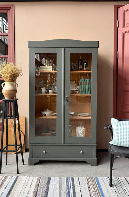 Ett gammalt vackert vitrinskåp med både runda och raka former i Dakaboms mörka grågröna färg. 