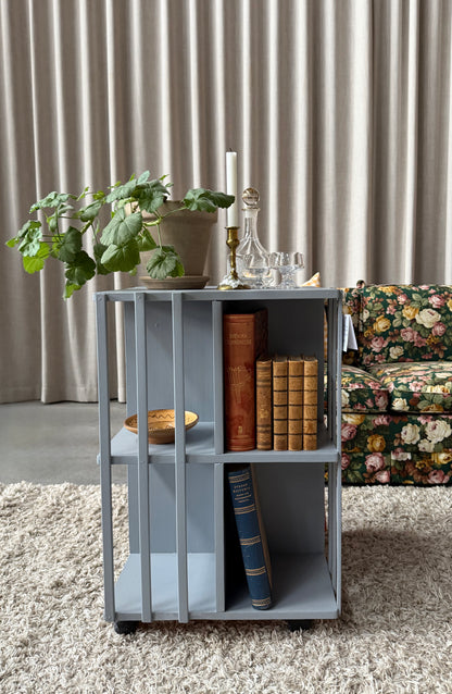 Ett vackert kvadratiskt sidebord/litet bord på hjul med ljuvliga små hyllor/fack på alla sidor vilket gör att det passar fint i rummet som sidebord att fylla med böcker och andra stilleben. Det är varsamt renoverat och målat i en ljust blå/grå kulör. 