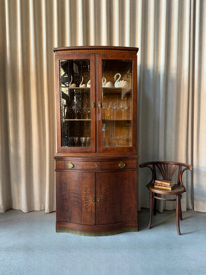 Ett elegant vitrinskåp i vacker Art Nouveau- stil med mjukt svängd front och utsmyckade detaljer som ger en tidlös och exklusiv känsla. Skåpet är tvådelat. Den övre vitrindelen med glasdörrar och hyllplan passar perfekt för att visa upp glas, porslin eller samlarföremål, medan den nedre delen är praktisk för förvaring. Skåpet har originalglas med dekorativ fasettslipning längs glasets kanter och tidstypiska slipade stjärnformade ornament. De mjuka formerna, gedigna hantverket och fantastiska detaljerna i vi