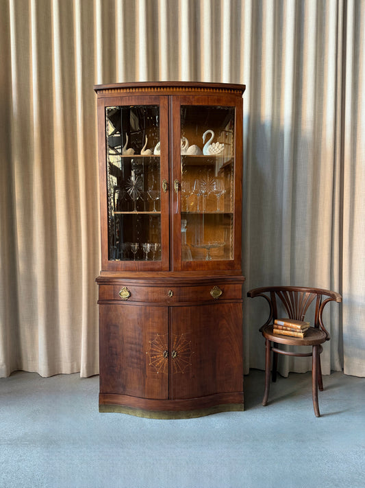 Ett elegant vitrinskåp i vacker Art Nouveau- stil med mjukt svängd front och utsmyckade detaljer som ger en tidlös och exklusiv känsla. Skåpet är tvådelat. Den övre vitrindelen med glasdörrar och hyllplan passar perfekt för att visa upp glas, porslin eller samlarföremål, medan den nedre delen är praktisk för förvaring. Skåpet har originalglas med dekorativ fasettslipning längs glasets kanter och tidstypiska slipade stjärnformade ornament. De mjuka formerna, gedigna hantverket och fantastiska detaljerna i vi