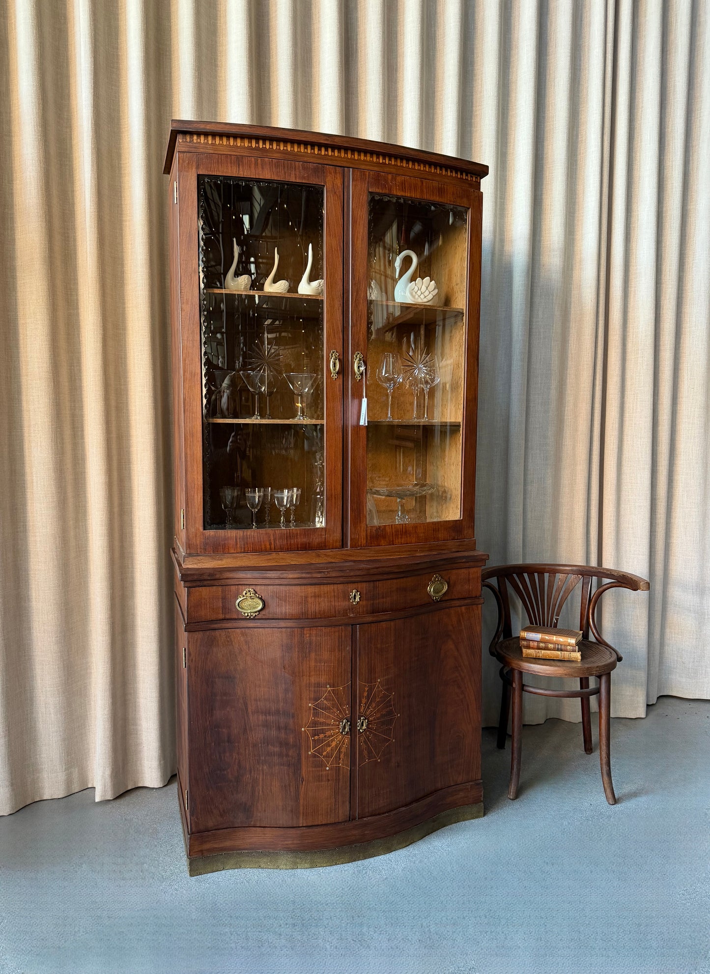 Ett elegant vitrinskåp i vacker Art Nouveau- stil med mjukt svängd front och utsmyckade detaljer som ger en tidlös och exklusiv känsla. Skåpet är tvådelat. Den övre vitrindelen med glasdörrar och hyllplan passar perfekt för att visa upp glas, porslin eller samlarföremål, medan den nedre delen är praktisk för förvaring. Skåpet har originalglas med dekorativ fasettslipning längs glasets kanter och tidstypiska slipade stjärnformade ornament. De mjuka formerna, gedigna hantverket och fantastiska detaljerna i vi