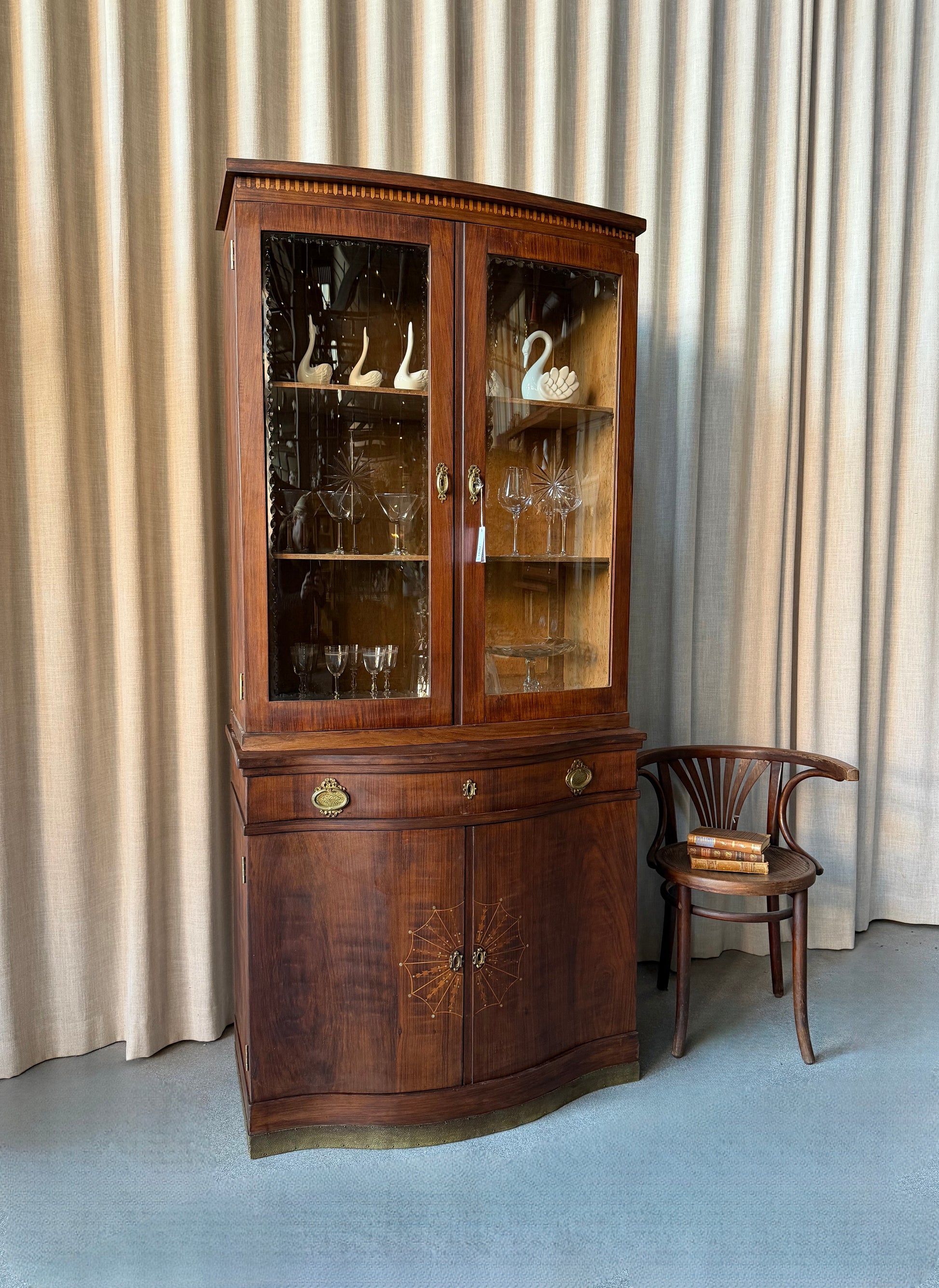 Ett elegant vitrinskåp i vacker Art Nouveau- stil med mjukt svängd front och utsmyckade detaljer som ger en tidlös och exklusiv känsla. Skåpet är tvådelat. Den övre vitrindelen med glasdörrar och hyllplan passar perfekt för att visa upp glas, porslin eller samlarföremål, medan den nedre delen är praktisk för förvaring. Skåpet har originalglas med dekorativ fasettslipning längs glasets kanter och tidstypiska slipade stjärnformade ornament. De mjuka formerna, gedigna hantverket och fantastiska detaljerna i vi
