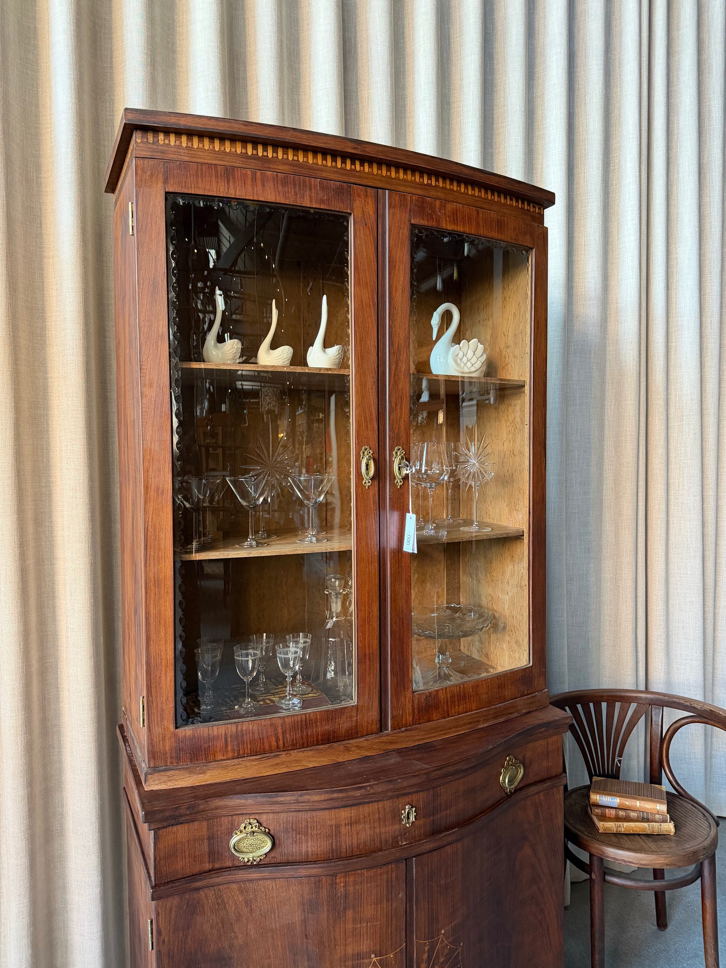 Ett elegant vitrinskåp i vacker Art Nouveau- stil med mjukt svängd front och utsmyckade detaljer som ger en tidlös och exklusiv känsla. Skåpet är tvådelat. Den övre vitrindelen med glasdörrar och hyllplan passar perfekt för att visa upp glas, porslin eller samlarföremål, medan den nedre delen är praktisk för förvaring. Skåpet har originalglas med dekorativ fasettslipning längs glasets kanter och tidstypiska slipade stjärnformade ornament. De mjuka formerna, gedigna hantverket och fantastiska detaljerna i vi