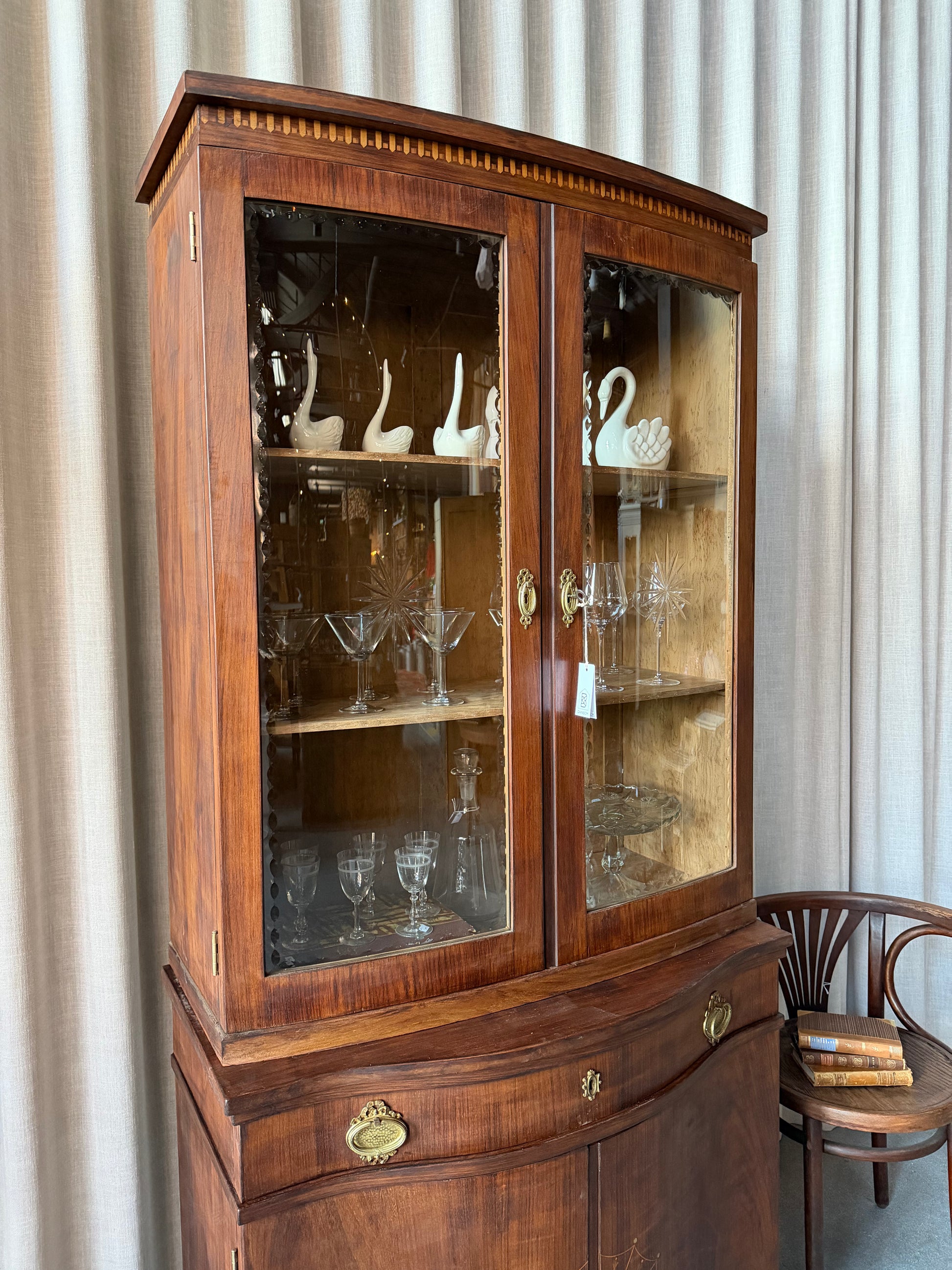 Ett elegant vitrinskåp i vacker Art Nouveau- stil med mjukt svängd front och utsmyckade detaljer som ger en tidlös och exklusiv känsla. Skåpet är tvådelat. Den övre vitrindelen med glasdörrar och hyllplan passar perfekt för att visa upp glas, porslin eller samlarföremål, medan den nedre delen är praktisk för förvaring. Skåpet har originalglas med dekorativ fasettslipning längs glasets kanter och tidstypiska slipade stjärnformade ornament. De mjuka formerna, gedigna hantverket och fantastiska detaljerna i vi