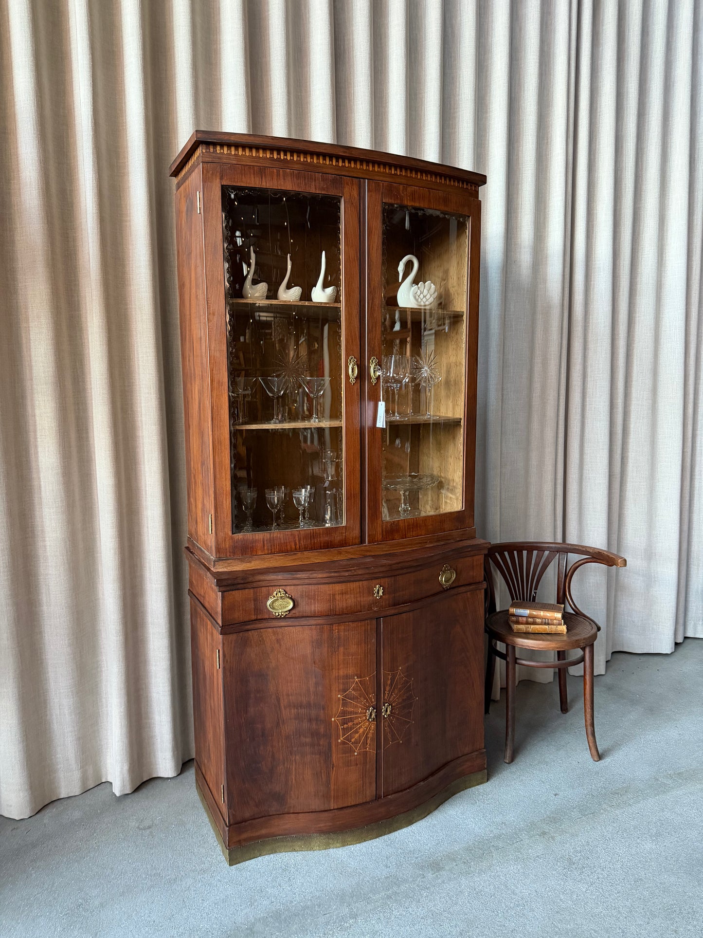 Ett elegant vitrinskåp i vacker Art Nouveau- stil med mjukt svängd front och utsmyckade detaljer som ger en tidlös och exklusiv känsla. Skåpet är tvådelat. Den övre vitrindelen med glasdörrar och hyllplan passar perfekt för att visa upp glas, porslin eller samlarföremål, medan den nedre delen är praktisk för förvaring. Skåpet har originalglas med dekorativ fasettslipning längs glasets kanter och tidstypiska slipade stjärnformade ornament. De mjuka formerna, gedigna hantverket och fantastiska detaljerna i vi