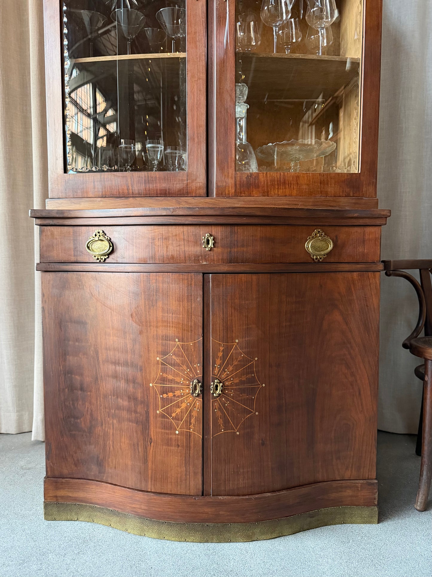 Ett elegant vitrinskåp i vacker Art Nouveau- stil med mjukt svängd front och utsmyckade detaljer som ger en tidlös och exklusiv känsla. Skåpet är tvådelat. Den övre vitrindelen med glasdörrar och hyllplan passar perfekt för att visa upp glas, porslin eller samlarföremål, medan den nedre delen är praktisk för förvaring. Skåpet har originalglas med dekorativ fasettslipning längs glasets kanter och tidstypiska slipade stjärnformade ornament. De mjuka formerna, gedigna hantverket och fantastiska detaljerna i vi
