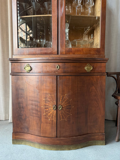 Ett elegant vitrinskåp i vacker Art Nouveau- stil med mjukt svängd front och utsmyckade detaljer som ger en tidlös och exklusiv känsla. Skåpet är tvådelat. Den övre vitrindelen med glasdörrar och hyllplan passar perfekt för att visa upp glas, porslin eller samlarföremål, medan den nedre delen är praktisk för förvaring. Skåpet har originalglas med dekorativ fasettslipning längs glasets kanter och tidstypiska slipade stjärnformade ornament. De mjuka formerna, gedigna hantverket och fantastiska detaljerna i vi