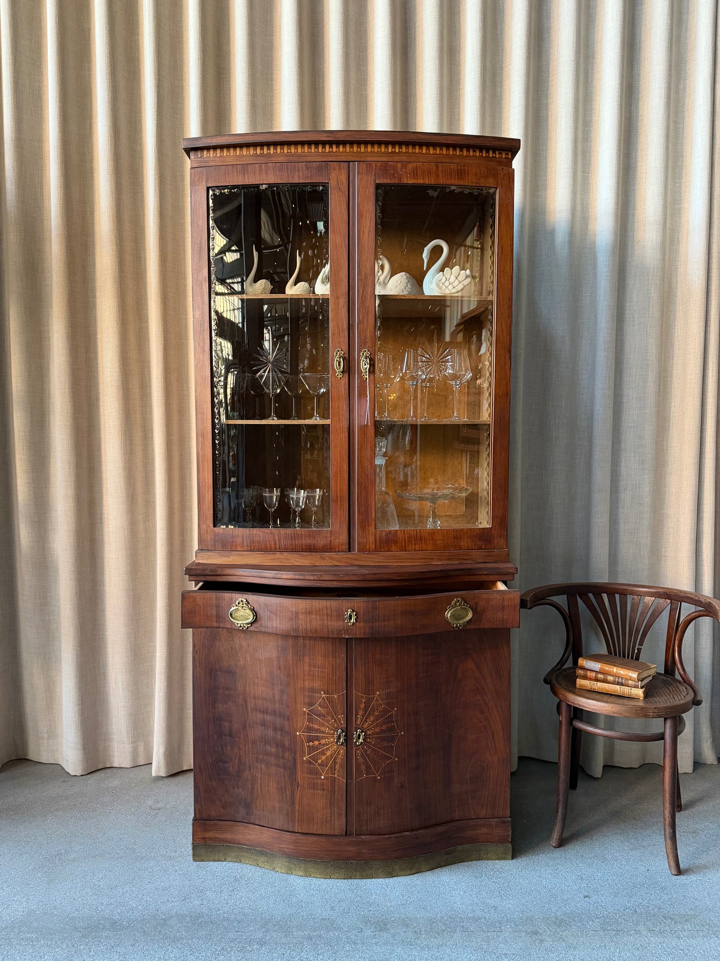 Ett elegant vitrinskåp i vacker Art Nouveau- stil med mjukt svängd front och utsmyckade detaljer som ger en tidlös och exklusiv känsla. Skåpet är tvådelat. Den övre vitrindelen med glasdörrar och hyllplan passar perfekt för att visa upp glas, porslin eller samlarföremål, medan den nedre delen är praktisk för förvaring. Skåpet har originalglas med dekorativ fasettslipning längs glasets kanter och tidstypiska slipade stjärnformade ornament. De mjuka formerna, gedigna hantverket och fantastiska detaljerna i vi