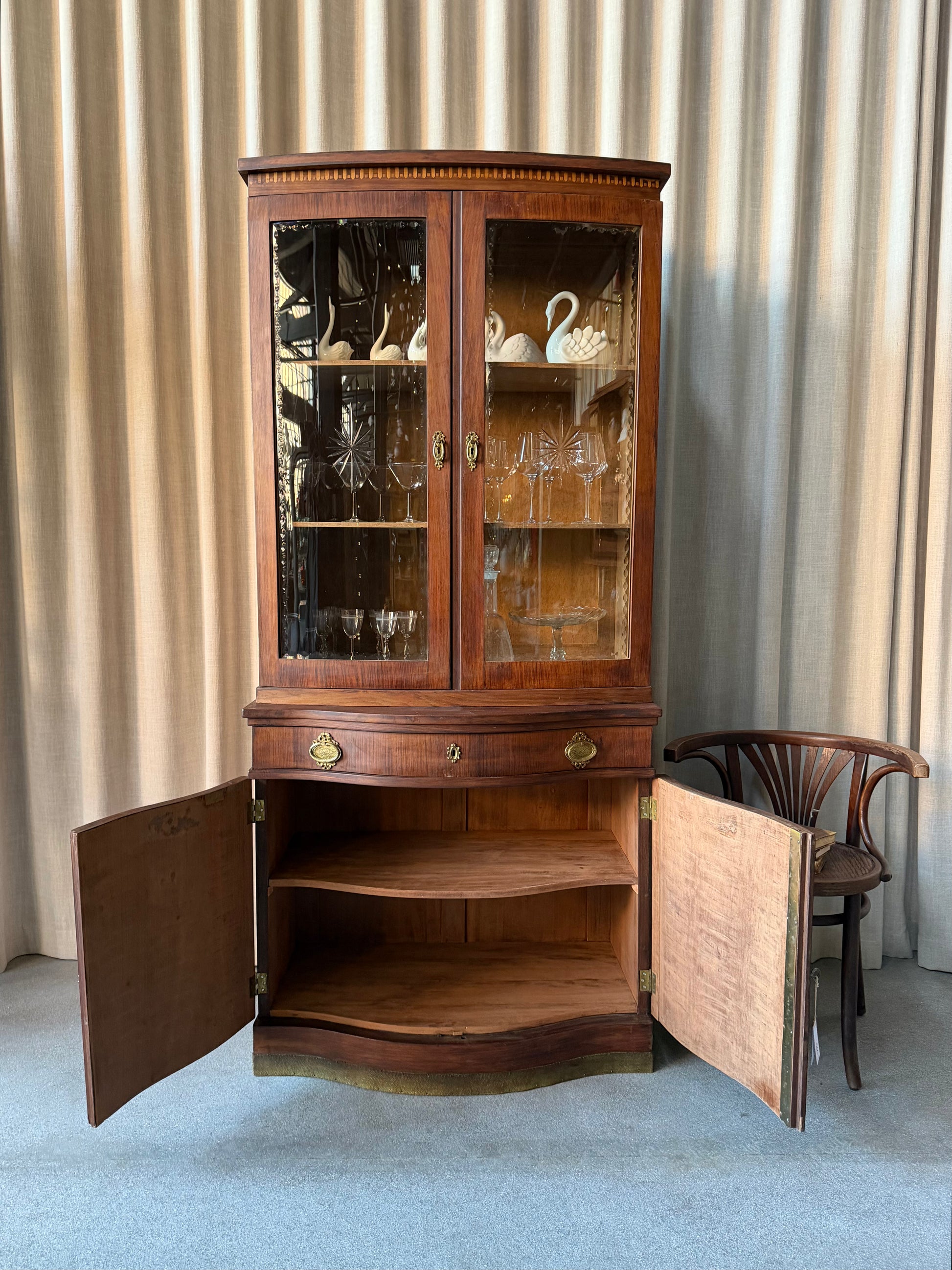 Ett elegant vitrinskåp i vacker Art Nouveau- stil med mjukt svängd front och utsmyckade detaljer som ger en tidlös och exklusiv känsla. Skåpet är tvådelat. Den övre vitrindelen med glasdörrar och hyllplan passar perfekt för att visa upp glas, porslin eller samlarföremål, medan den nedre delen är praktisk för förvaring. Skåpet har originalglas med dekorativ fasettslipning längs glasets kanter och tidstypiska slipade stjärnformade ornament. De mjuka formerna, gedigna hantverket och fantastiska detaljerna i vi