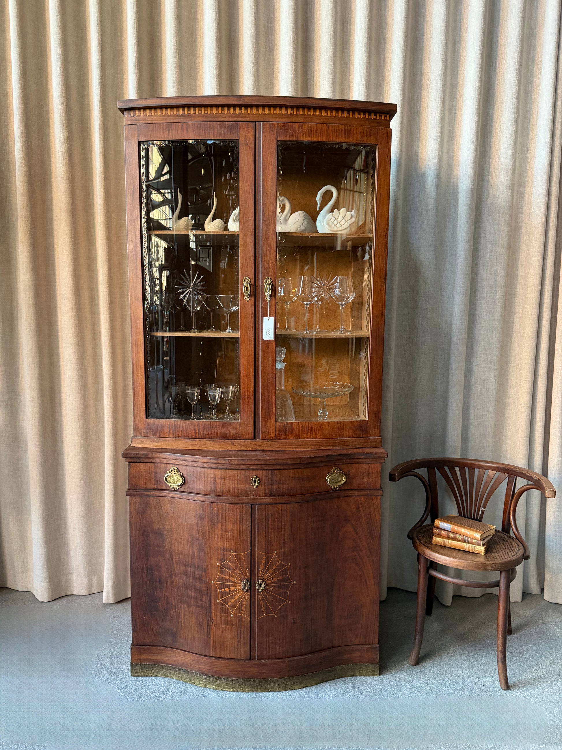 Ett elegant vitrinskåp i vacker Art Nouveau- stil med mjukt svängd front och utsmyckade detaljer som ger en tidlös och exklusiv känsla. Skåpet är tvådelat. Den övre vitrindelen med glasdörrar och hyllplan passar perfekt för att visa upp glas, porslin eller samlarföremål, medan den nedre delen är praktisk för förvaring. Skåpet har originalglas med dekorativ fasettslipning längs glasets kanter och tidstypiska slipade stjärnformade ornament. De mjuka formerna, gedigna hantverket och fantastiska detaljerna i vi