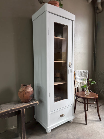 Smalt vitrinskåp med en enkeldörr som är vänsterhängd. Skåpet är vitt med glas i dörren och har en låda längst ner.  Till höger står en stol och till vänster en bänk med en urna på. Bilden är tagen snett från sidan.