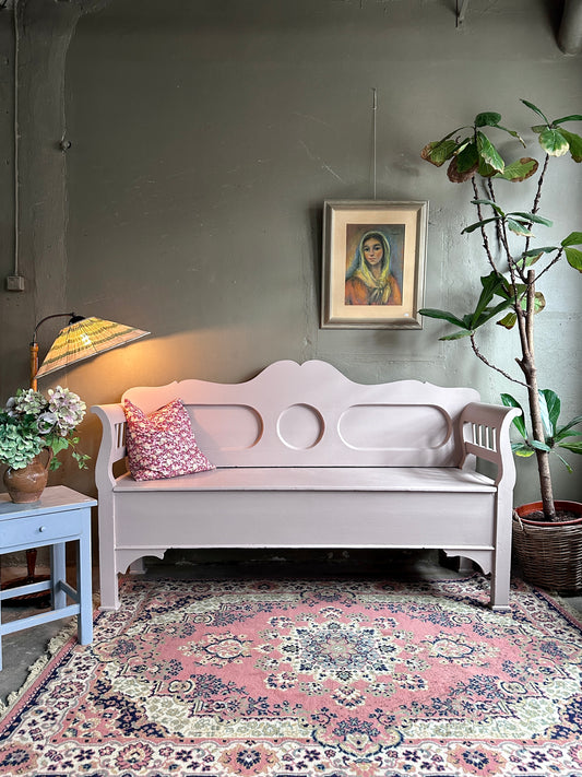 Kökssoffa i ljust rosa kulör. På bilden syns också en matta, pall, golvlampa, växt samt en tavla. Bilden är tagen rakt framifrån.