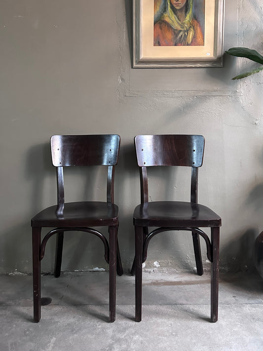 2 styck stolar i mörkt trä. Bilden är tagen rakt framifrån. På golvet står en urna och på väggen hänger en tavla. 