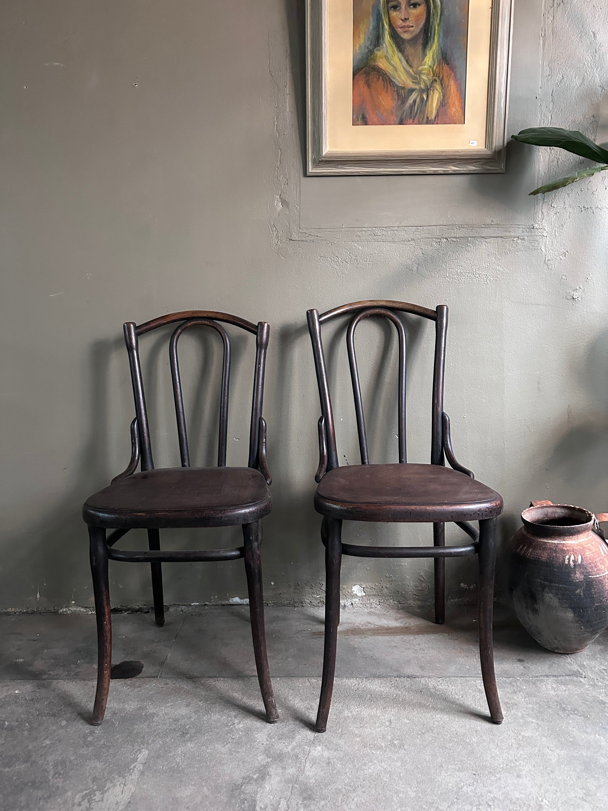 2 st gamla stolar i mörkt trä. Bilden är tagen rakt framifrån. På golvet  bredvid står en urna och en tavla hänger på väggen. 