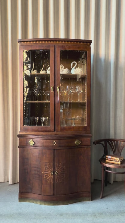 Ett elegant vitrinskåp i vacker Art Nouveau- stil med mjukt svängd front och utsmyckade detaljer som ger en tidlös och exklusiv känsla. Skåpet är tvådelat. Den övre vitrindelen med glasdörrar och hyllplan passar perfekt för att visa upp glas, porslin eller samlarföremål, medan den nedre delen är praktisk för förvaring. Skåpet har originalglas med dekorativ fasettslipning längs glasets kanter och tidstypiska slipade stjärnformade ornament. De mjuka formerna, gedigna hantverket och fantastiska detaljerna i vi