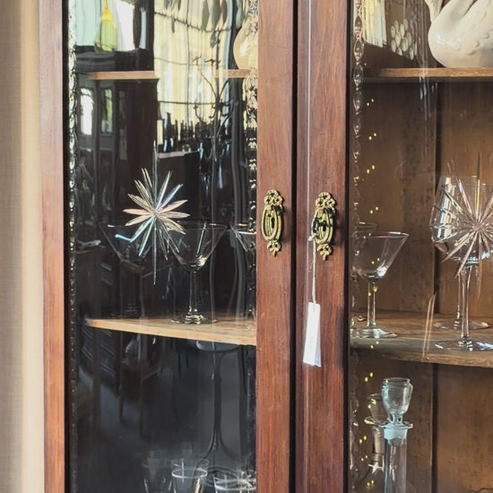 Ett elegant vitrinskåp i vacker Art Nouveau- stil med mjukt svängd front och utsmyckade detaljer som ger en tidlös och exklusiv känsla. Skåpet är tvådelat. Den övre vitrindelen med glasdörrar och hyllplan passar perfekt för att visa upp glas, porslin eller samlarföremål, medan den nedre delen är praktisk för förvaring. Skåpet har originalglas med dekorativ fasettslipning längs glasets kanter och tidstypiska slipade stjärnformade ornament. De mjuka formerna, gedigna hantverket och fantastiska detaljerna i vi