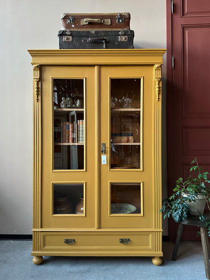 Ett gammalt, otroligt vackert vitrinskåp i Dakaboms ockragula kulör med fina detaljer i de raka formerna och det arbetade krönet. Skåpet har i dagsläget 3 st nya och fräscha hyllplan. 
Det har dessutom en bred praktisk låda längst ner. 