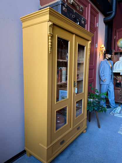 Ett gammalt, otroligt vackert vitrinskåp i Dakaboms ockragula kulör med fina detaljer i de raka formerna och det arbetade krönet. Skåpet har i dagsläget 3 st nya och fräscha hyllplan. 
Det har dessutom en bred praktisk låda längst ner. 