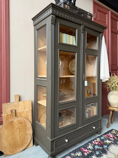 Ett gammalt, fantastiskt vackert vitrinskåp med fina detaljer i de raka formerna och det arbetade krönet. Skåpet har i dagsläget 3 st nya och fräscha hyllplan.Det har dessutom en bred praktisk låda längst ner med vackra porslinsknoppar. Skåpet är varsamt renoverat och slutligen penselmålat med en matt, vattenbaserad snickerifärg i en brun/grå kulör. 