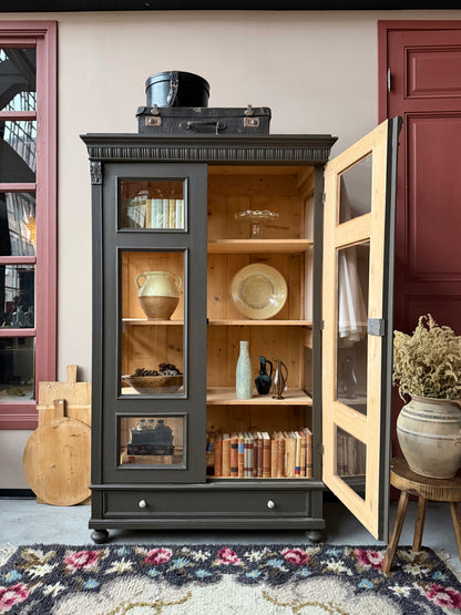 Ett gammalt, fantastiskt vackert vitrinskåp med fina detaljer i de raka formerna och det arbetade krönet. Skåpet har i dagsläget 3 st nya och fräscha hyllplan.Det har dessutom en bred praktisk låda längst ner med vackra porslinsknoppar. Skåpet är varsamt renoverat och slutligen penselmålat med en matt, vattenbaserad snickerifärg i en brun/grå kulör. 